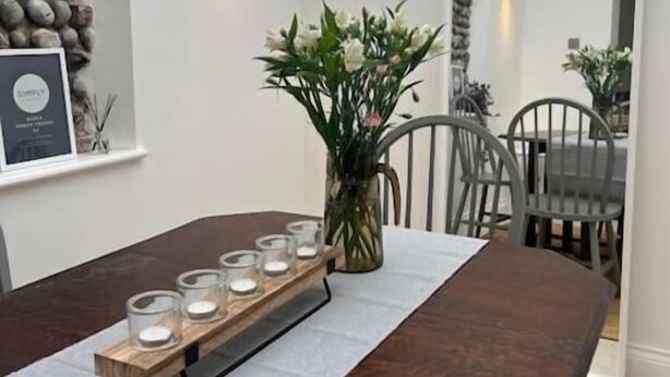 A modern dining area with a long wooden table, candle holders, and a vase of flowers. - Samphire House (Cromer)