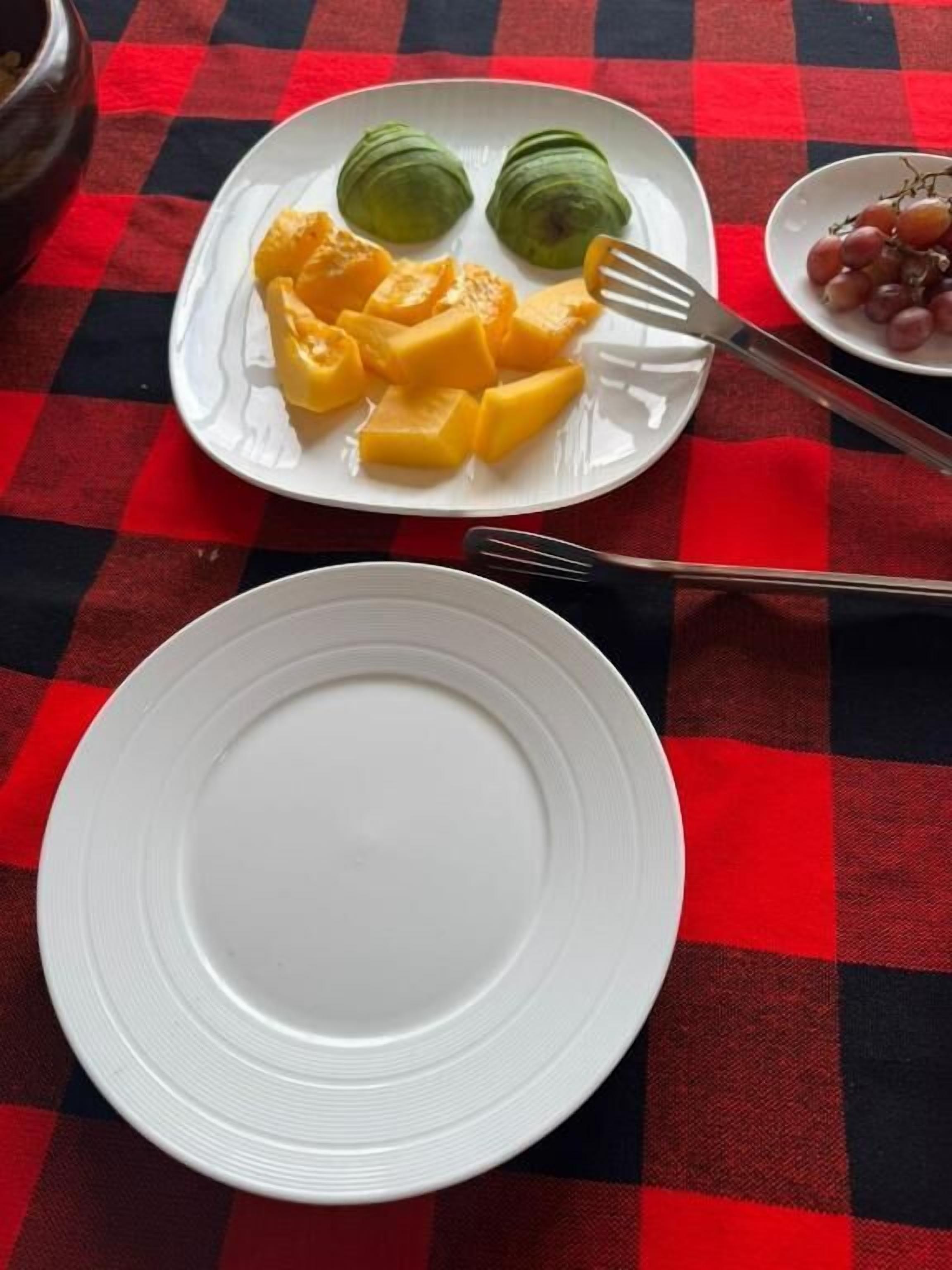 A table with a red and black checkered tablecloth, a white plate with mango chunks and two green rolls, and a bowl of grapes.