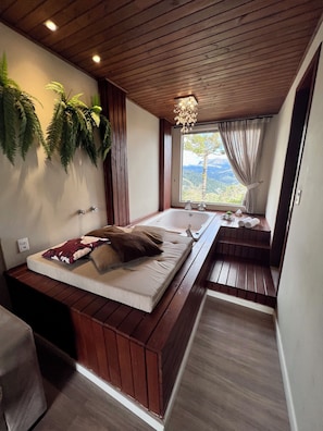 A bedroom with a wooden ceiling, a large window with curtains, a bed with pillows, and a wooden bench by the window. - Pousada Hinckel (Alfredo Wagner)