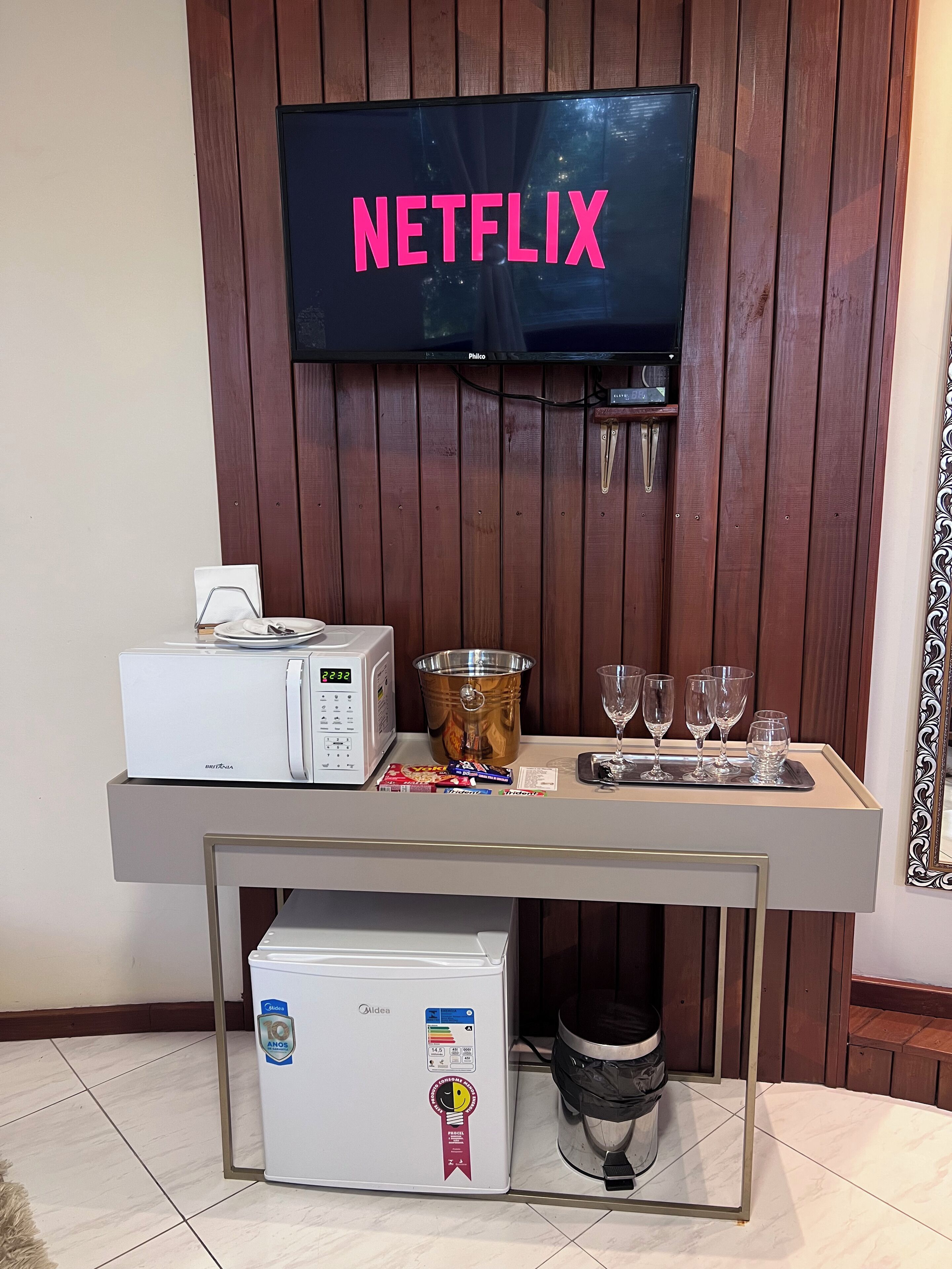 Quarto com parede de madeira, uma televisão exibindo "NETFLIX", um minibar, um pequeno frigorífico e uma lixeira.