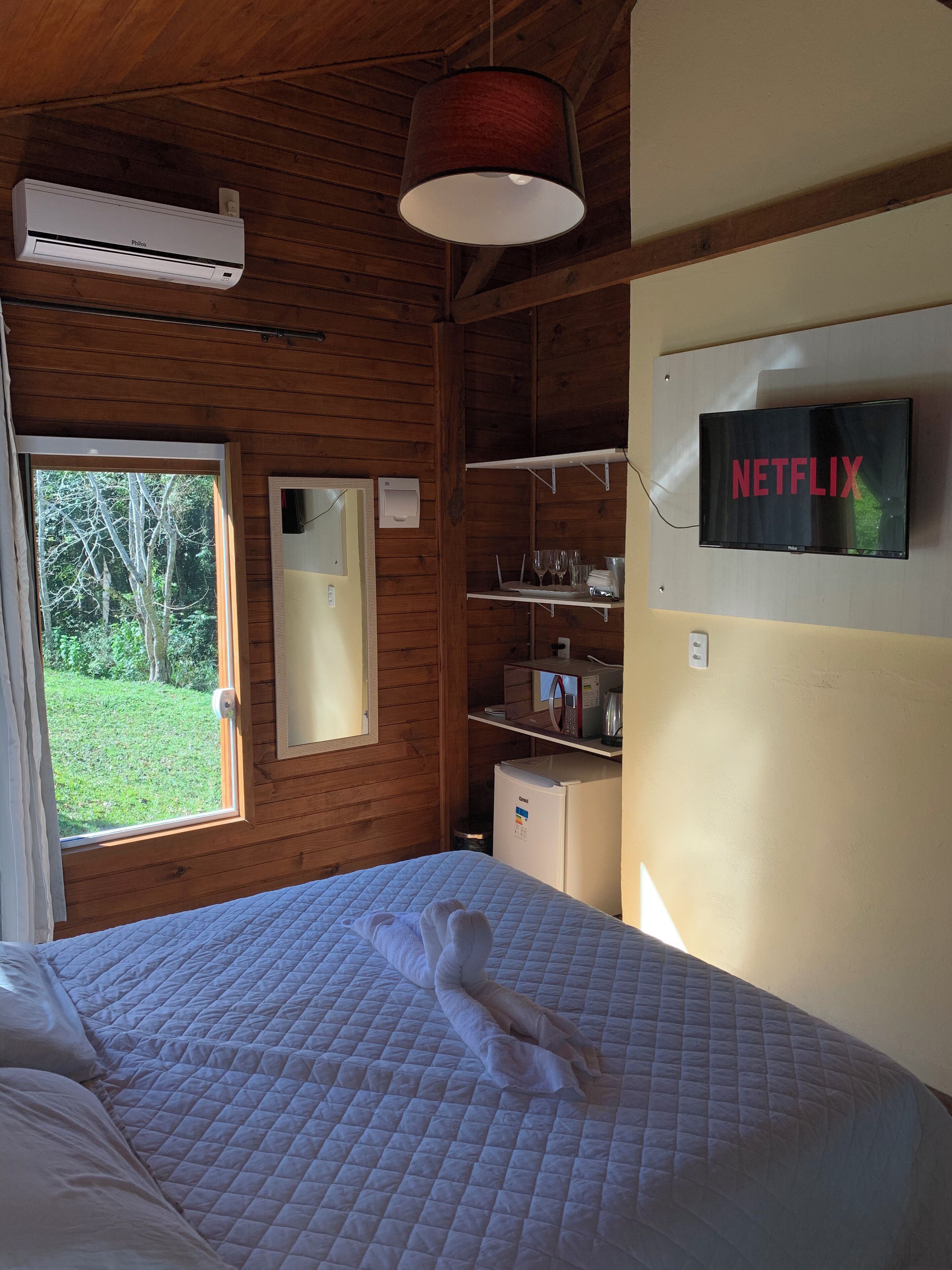 A wooden cabin room with a bed, a mounted TV displaying 'NETFLIX', a window, and a kitchenette area.