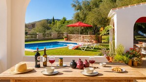 On voit une piscine avec des chaises longues et un parasol rouge, une table avec des bouteilles de vin et des verres, ainsi que diverses plantes en pot.