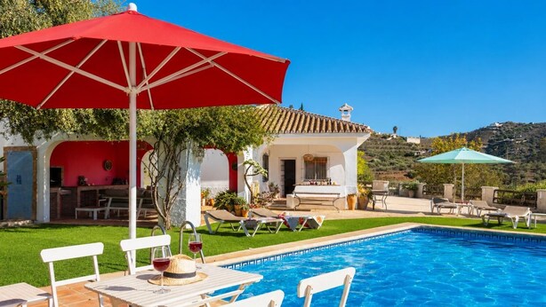 Un espace piscine avec des chaises longues et une table, un parasol rouge et un bâtiment doté d’un toit en tuiles.