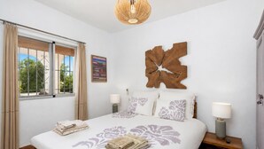 Une chambre à coucher avec un grand lit, deux tables de chevet, une œuvre d’art accrochée au mur, une fenêtre avec des rideaux et un luminaire suspendu.