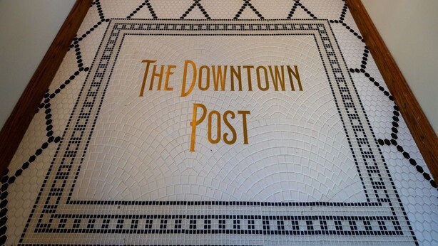 A hallway with a patterned floor leading to a door, a staircase, and a wall-mounted television. - The Downtown Post (Elizabethton)