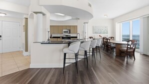 A modern kitchen with a curved island, bar stools, and a dining area with a large table and chairs. - Salida del Sol 1101 (Daytona Beach Shores)