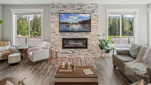 A modern living room with a stone wall, a flat-screen TV, a fireplace, and large windows with a view of a lake and forest. - Waterfront home w/River access & Amazing views (Post Falls)