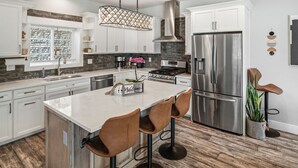 A modern kitchen with white cabinets, a stainless steel refrigerator, a large island with bar stools, and a hanging light fixture. - Waterfront home w/River access & Amazing views (Post Falls)