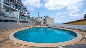 A swimming pool with a tiled deck, surrounded by a building with balconies and a view of the sea. - Amethyst by LovelyStay (Canico)
