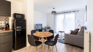 A modern living room with a grey sofa, a dining table with four chairs, and a kitchen area with a black refrigerator and microwave.
