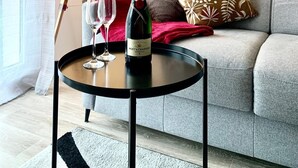 A modern living room with a grey sofa, a small round table with a bottle of champagne and two glasses, a floor lamp, and a decorative pillow.