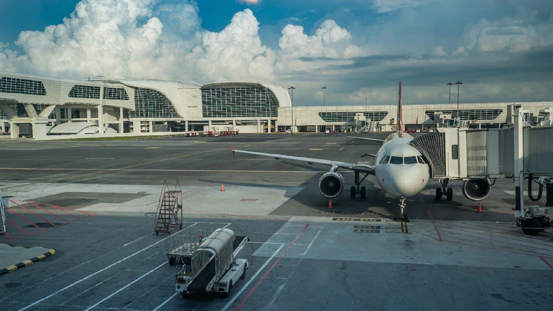 An airport terminal with a parked airplane connected to a jet bridge, a luggage conveyor, and a jet bridge in the background.