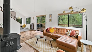 Una sala de estar moderna con sofá, mesa de centro y chimenea. Se ve una escalera que sube a un nivel superior, y un ventanal con vistas a árboles.