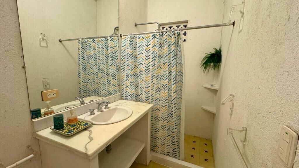 A bathroom with a white sink, a mirror, a shower with a patterned curtain, and a small shelf with toiletries.