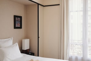 A hotel room with a bed, a nightstand, a framed picture, a window with curtains, and a view of buildings.