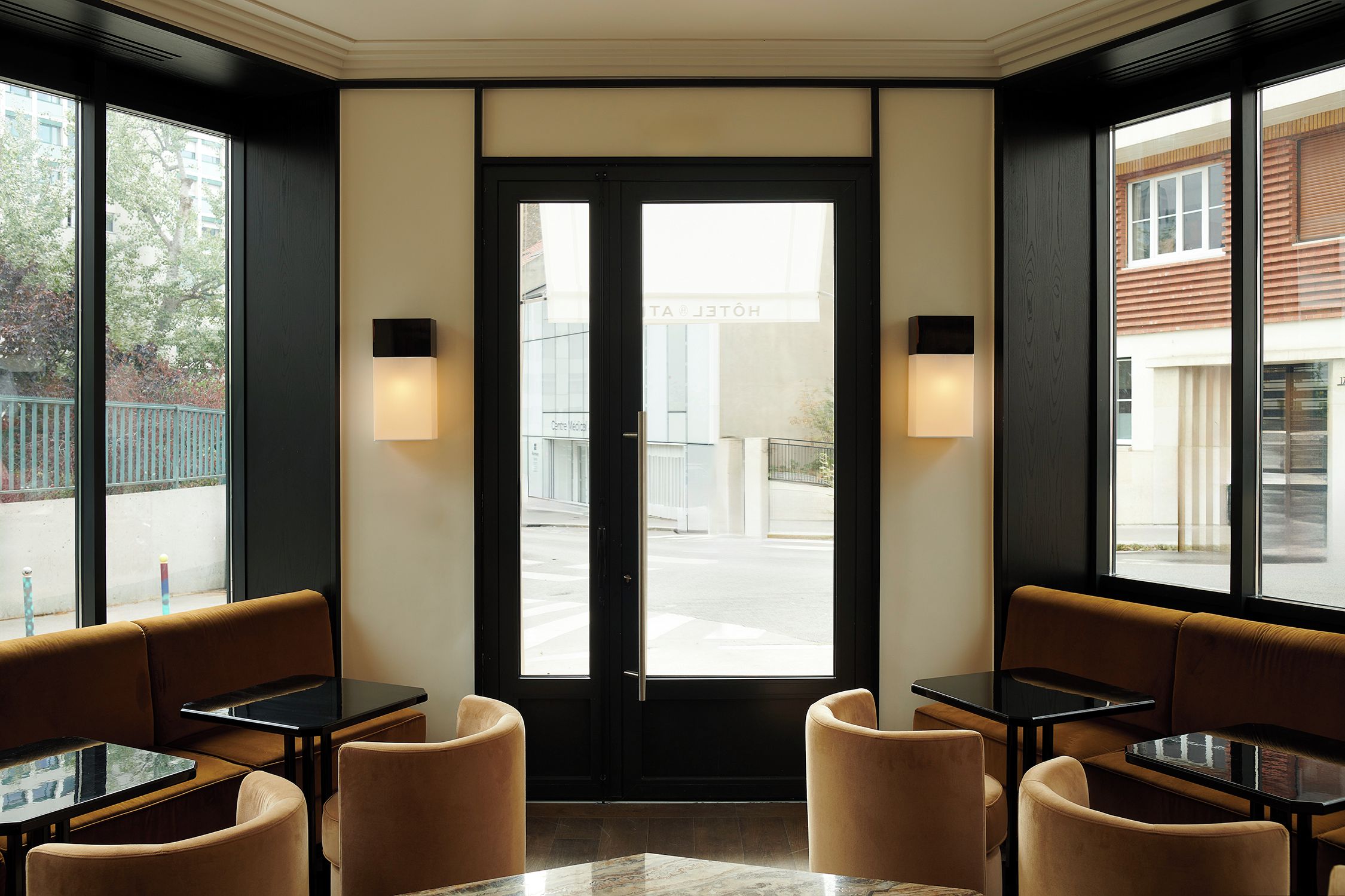 A hotel lobby with a double door entrance, seating area with tables and chairs, and large windows providing views of the street.