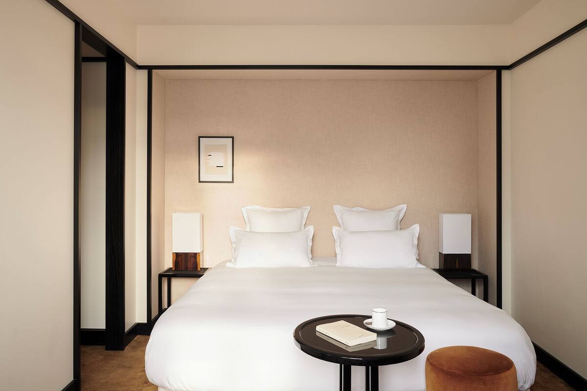 Quarto de hotel com uma cama grande, duas mesas de cabeceira, uma mesinha redonda com um livro e uma chávena, e um banquinho marrom.