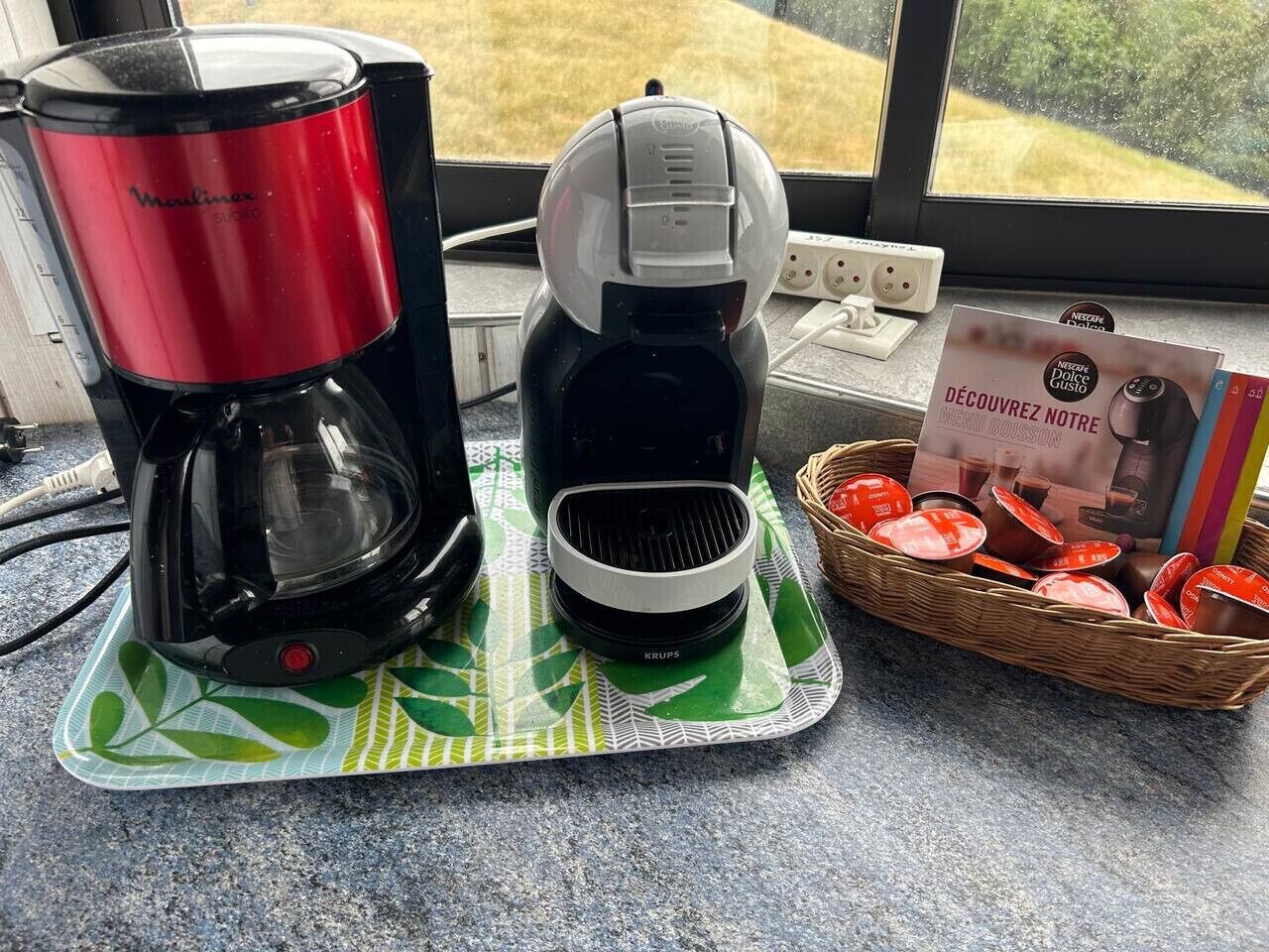 Une machine à café, une bouilloire et un panier de dosettes de café sur un plan de travail de cuisine.