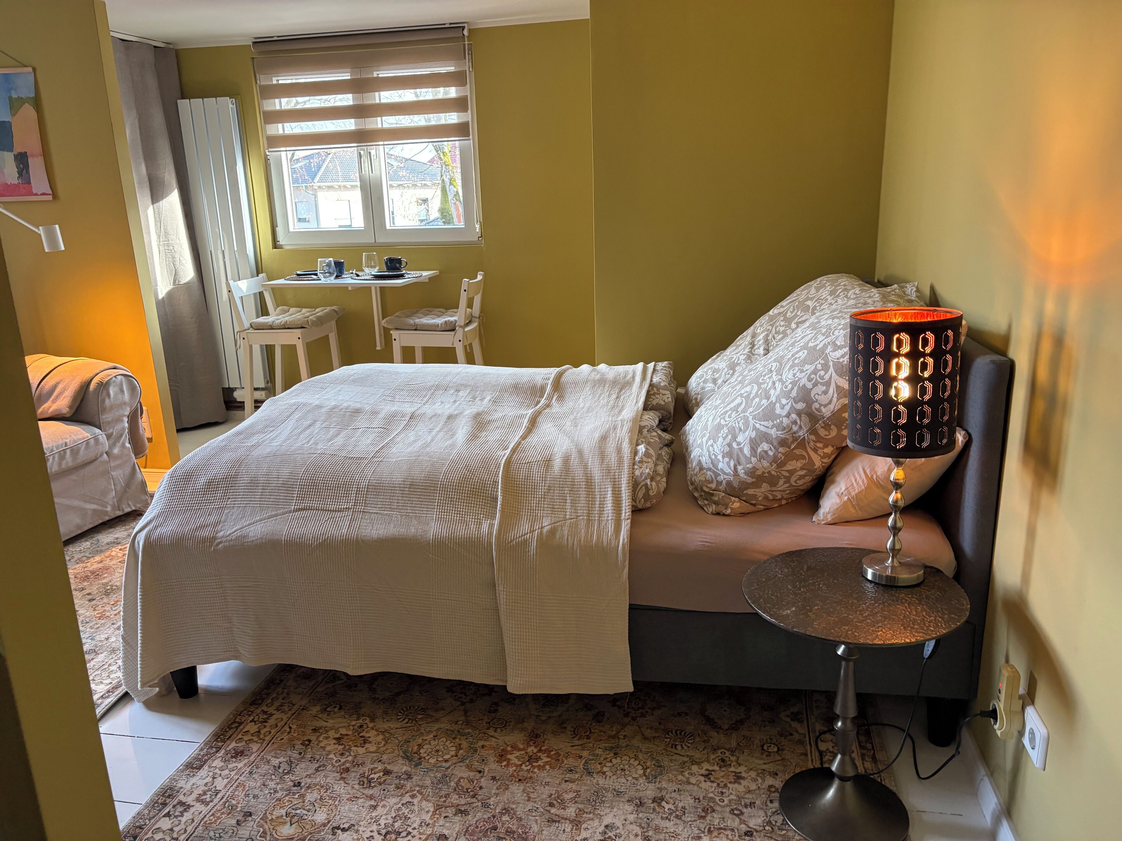 A bedroom with a bed, a bedside lamp, a small round table, and a window with blinds.