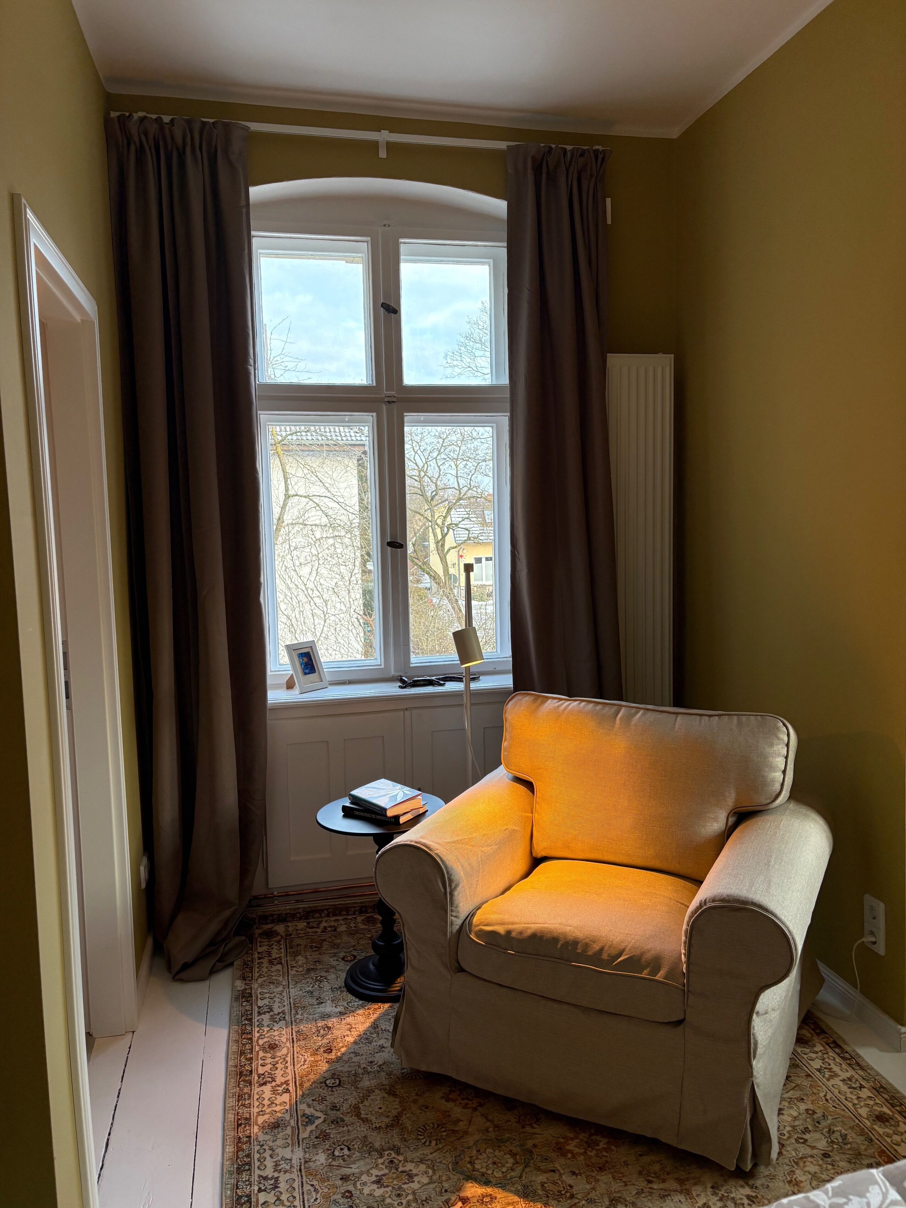 Ein Zimmer mit einem beigefarbenen Sessel, einem kleinen Tisch mit Büchern, einer Lampe und einem mit Vorhängen drapierten Fenster.