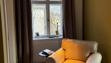 Ein Zimmer mit einem beigefarbenen Sessel, einem kleinen Tisch mit Büchern, einer Lampe und einem mit Vorhängen drapierten Fenster.