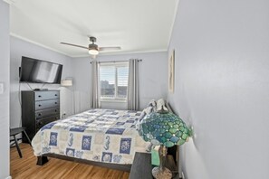 Ein Schlafzimmer mit Bett, Deckenventilator, Fernseher und einem Fenster mit Jalousien.