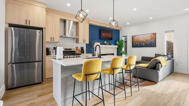 A modern kitchen with a central island, stainless steel appliances, and a dining area with yellow chairs. - New 1500 ft² Home w/ AC & Garage | 7 mins to DT (Calgary)