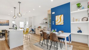 A modern kitchen with a central island, a dining area with chairs, and a blue accent wall. - New 1500 ft² Home w/ AC & Garage | 7 mins to DT (Calgary)