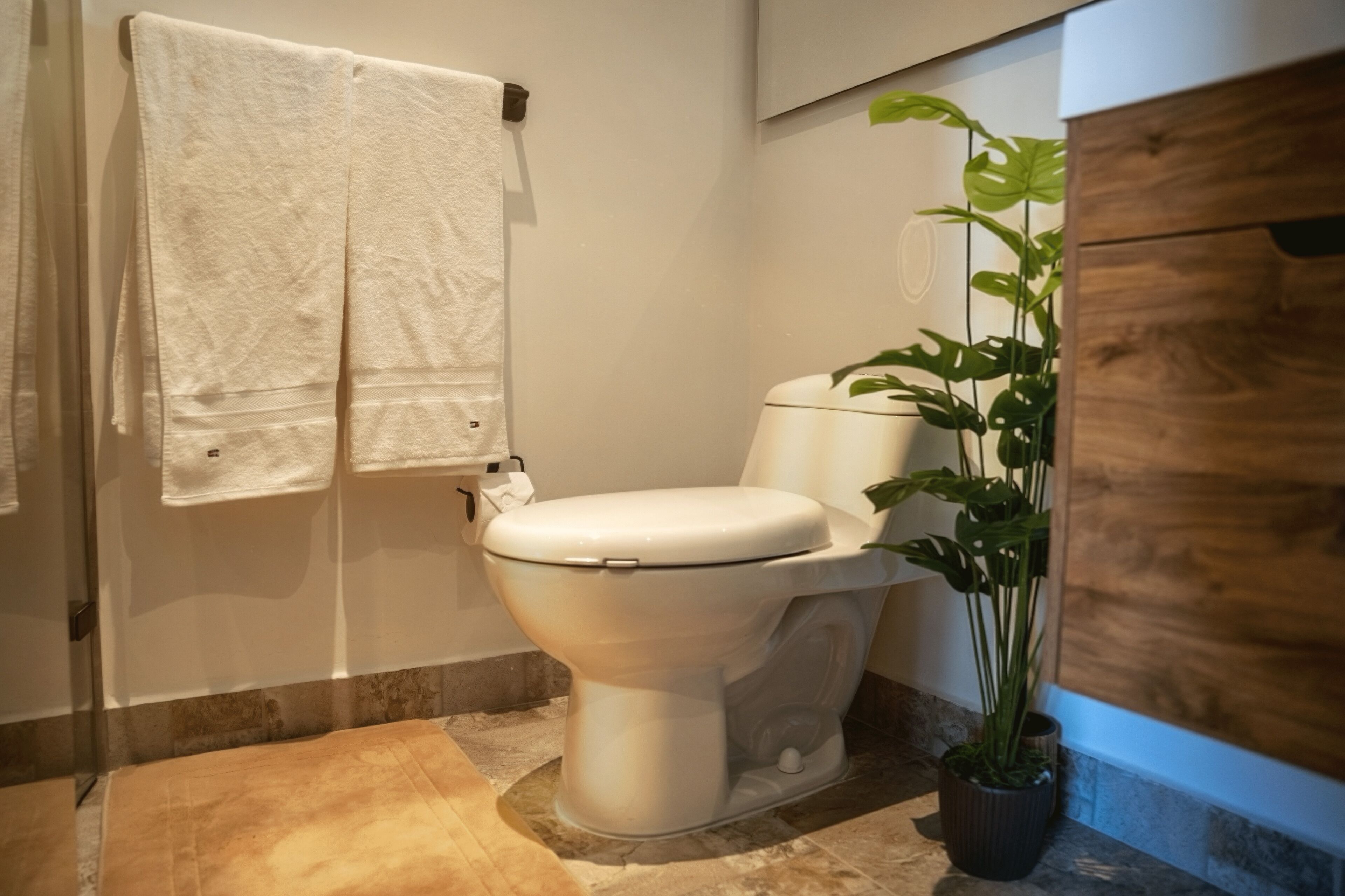 A modern bathroom with a white toilet, a towel rack with two towels, a potted plant, and a wooden cabinet.