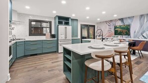A modern kitchen with teal cabinetry, a white island with bar stools, and a dining area with a table and chairs. - Pickleball-Basketball-Pool-HotTub-GameRoom-FirePit (Columbus)