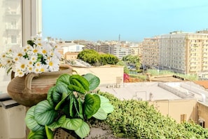 A balcony with a potted plant and flowers, overlooking a cityscape with buildings and a clear sky. - Il Panoramico Rooms (Cagliari)