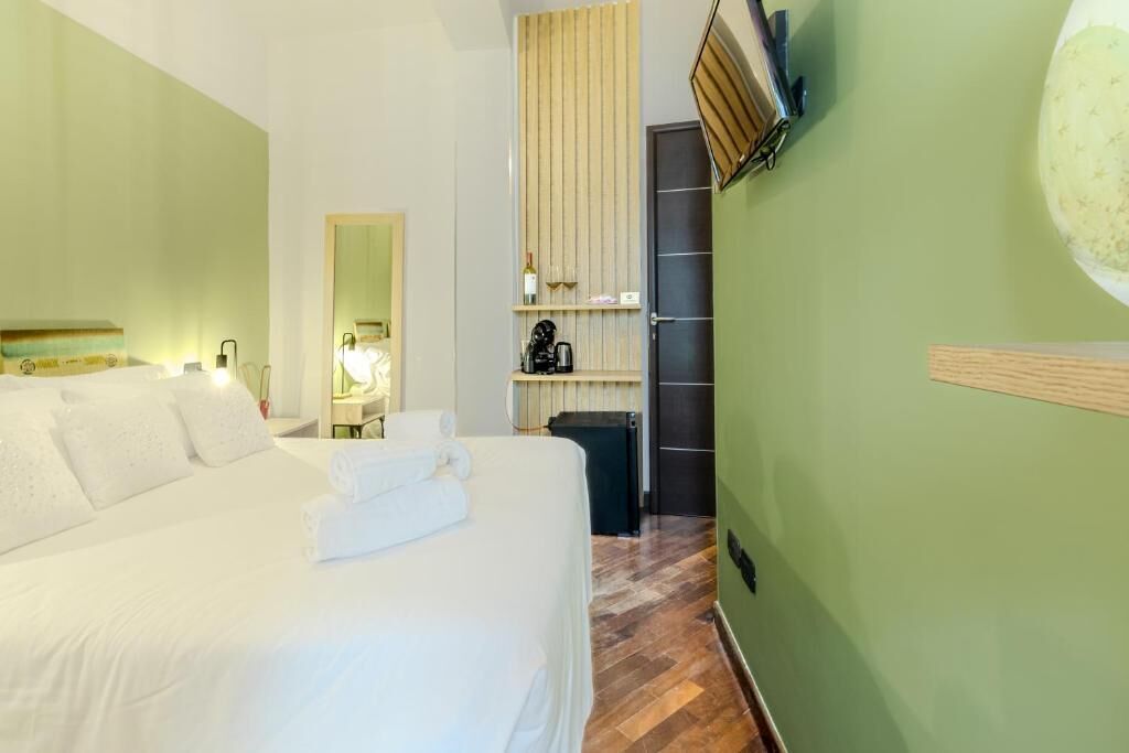 A hotel room with a large bed, a wooden floor, a green wall, a mirror, a shelf with a bottle and a kettle, and a door.