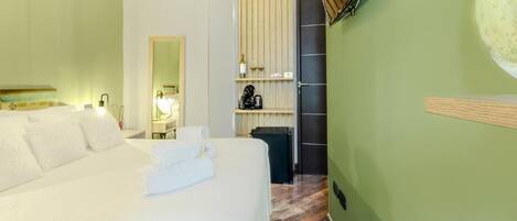 A hotel room with a large bed, a wooden floor, a green wall, a mirror, a shelf with a bottle and a kettle, and a door.