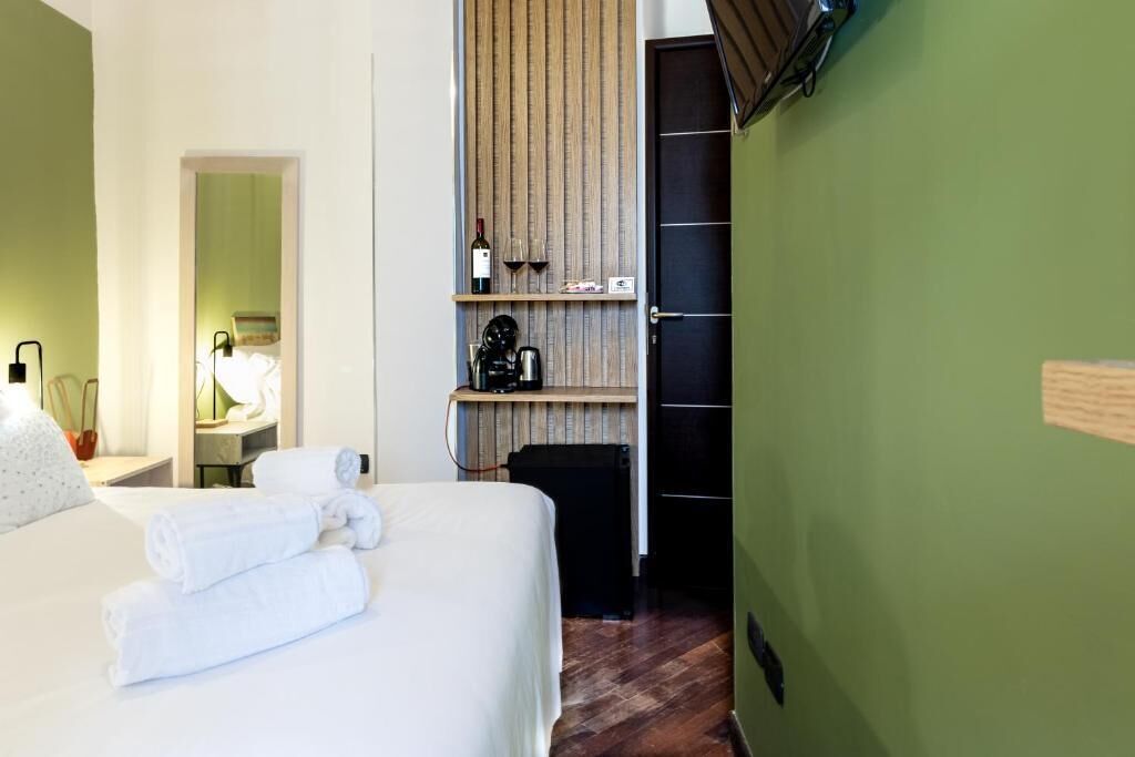 Habitación de hotel con cama, repisa de madera con una botella y una cafetera, y pared verde.