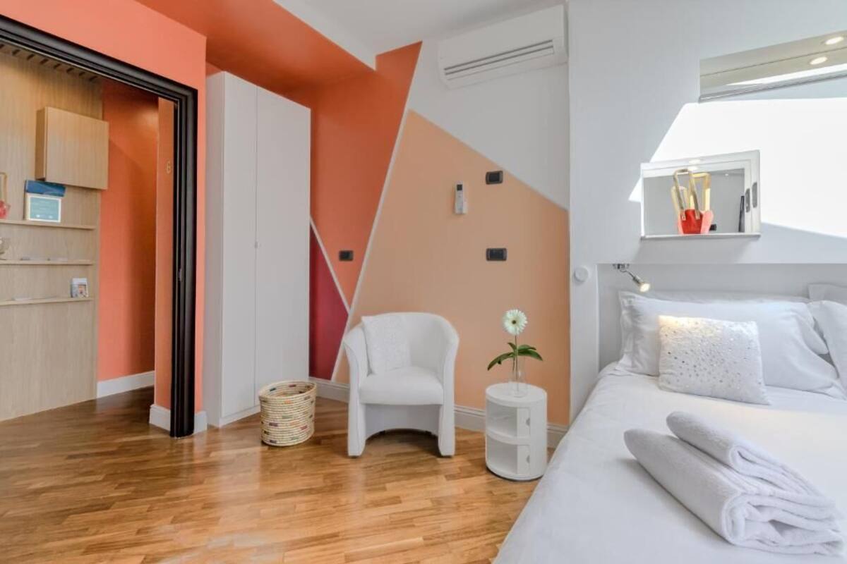 Une chambre d’hôtel moderne avec un plancher en bois, un lit blanc avec une literie blanche, une chaise blanche, une petite table d’appoint avec un vase, et un grand miroir au-dessus du lit.