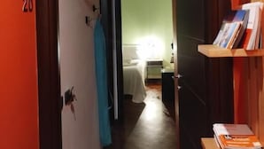 Een hal met een deur waarop '20' staat, een houten plank met boeken, en een bed in de aangrenzende kamer.