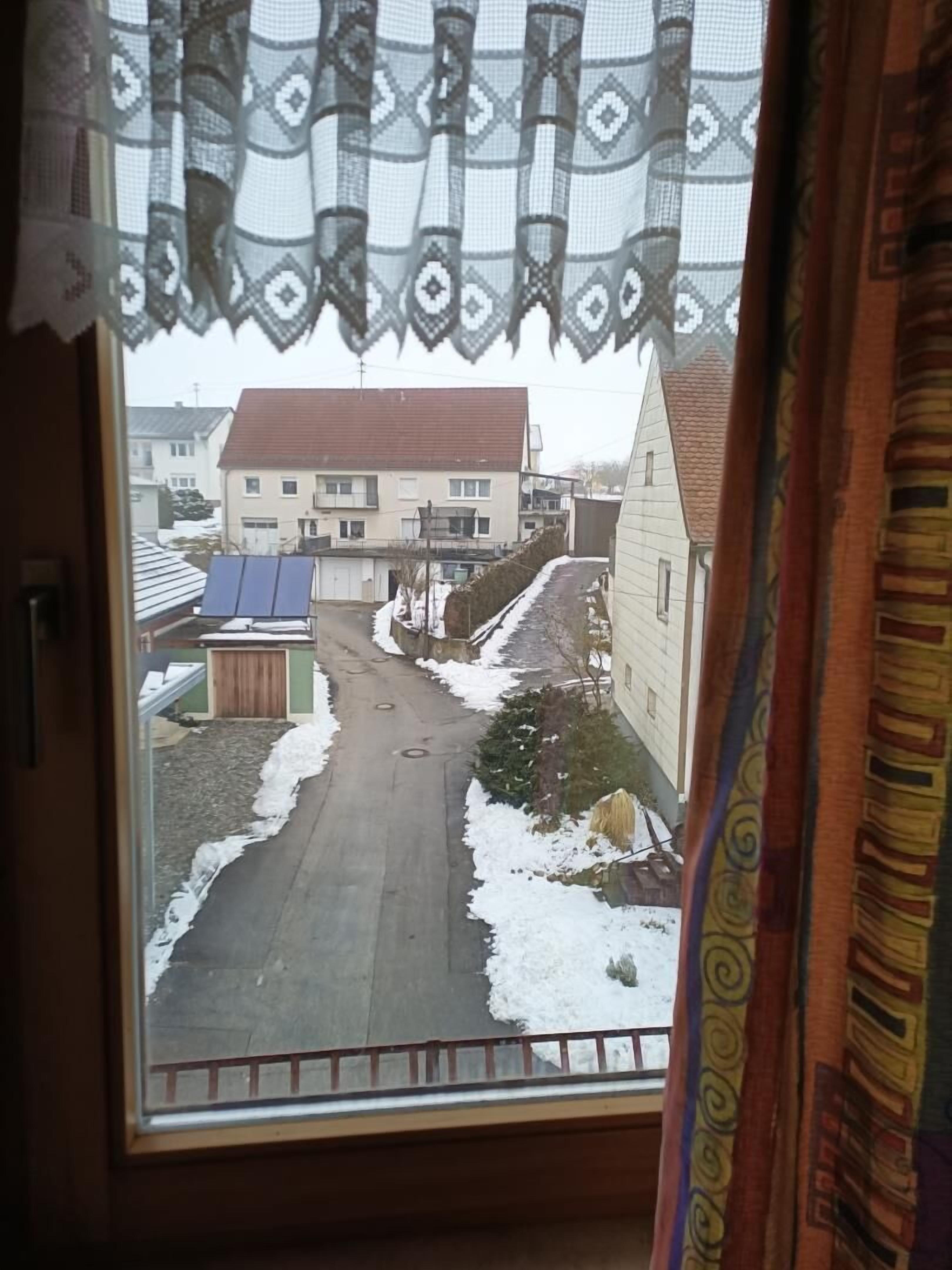 Blick aus dem Fenster auf eine Wohnstraße mit Häusern, einer asphaltierten Straße und Solaranlage auf einem Dach.