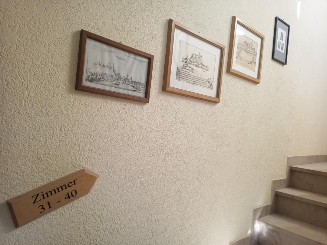 Gerahmte architektonische Zeichnungen an einer strukturierten Wand, ein Holzschild mit Zimmernummern und eine Treppe.