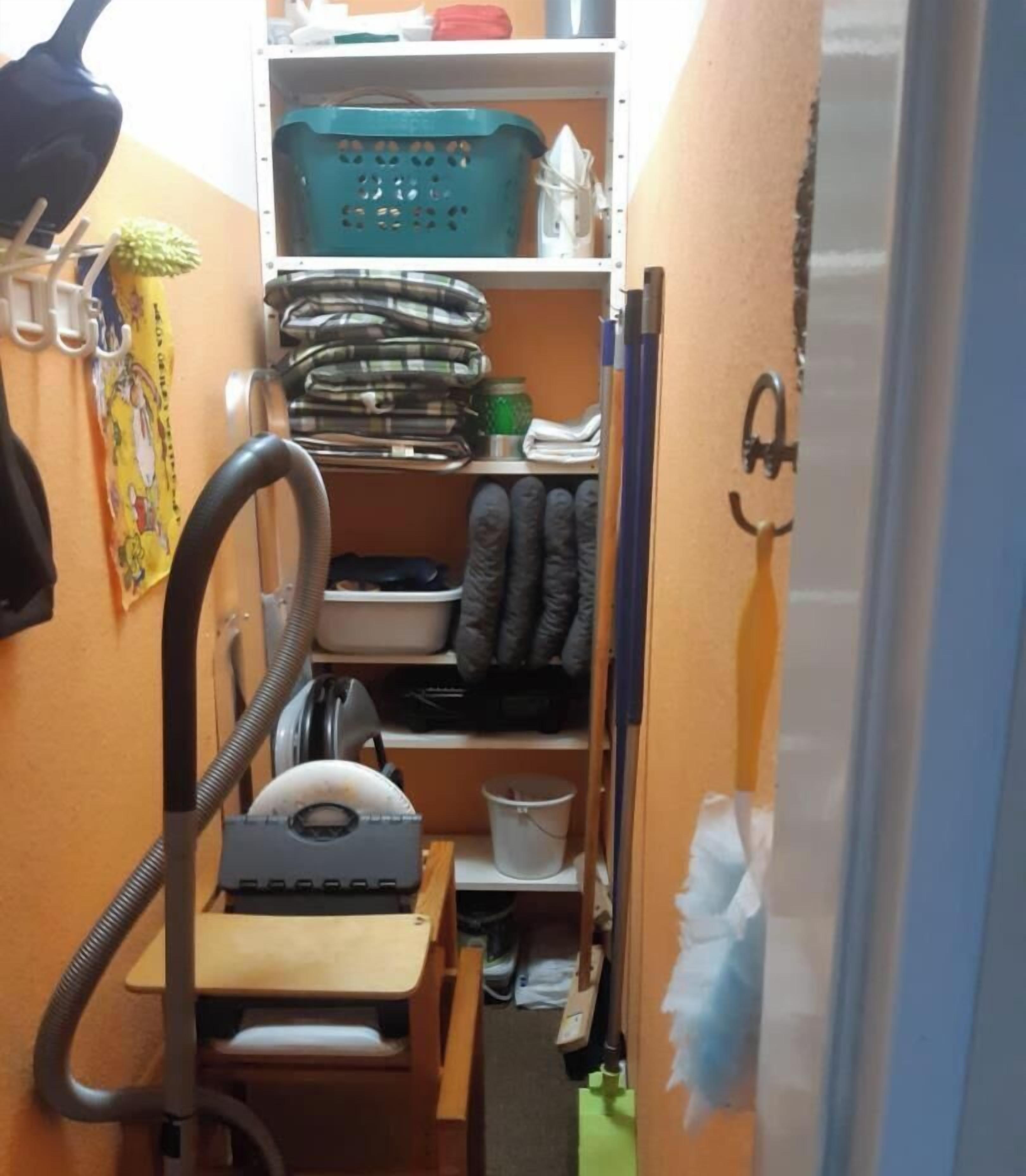 A narrow hallway with a vacuum cleaner, a shelf with various items, and a door with a smiley face sticker.