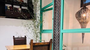 Un comedor con mesa y sillas de madera, piso con estampado verde, una estantería y una ventana con una lámpara decorativa.