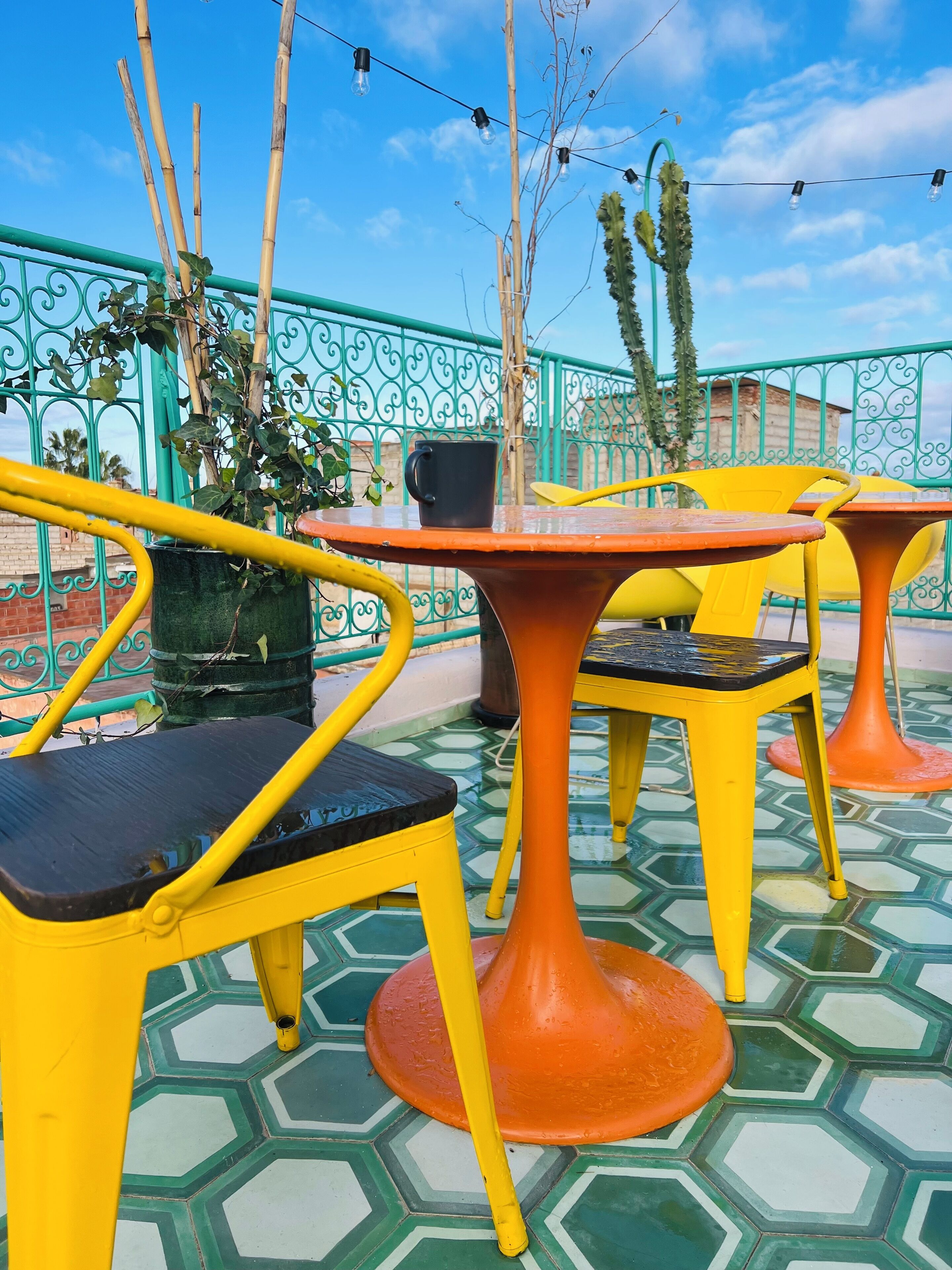 Outdoor seating with yellow chairs and orange tables, surrounded by a green fence and plants.