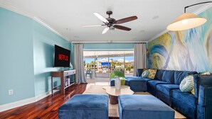 Uma sala de estar com um sofá azul, pufes, uma mesa de centro e uma TV exibindo 'NETFLIX'.