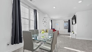 A modern dining area with a glass table and cushioned chairs, a red sofa, and a TV mounted on the wall. - Charming 3-bedroom Home in Southwest Detroit (Detroit)