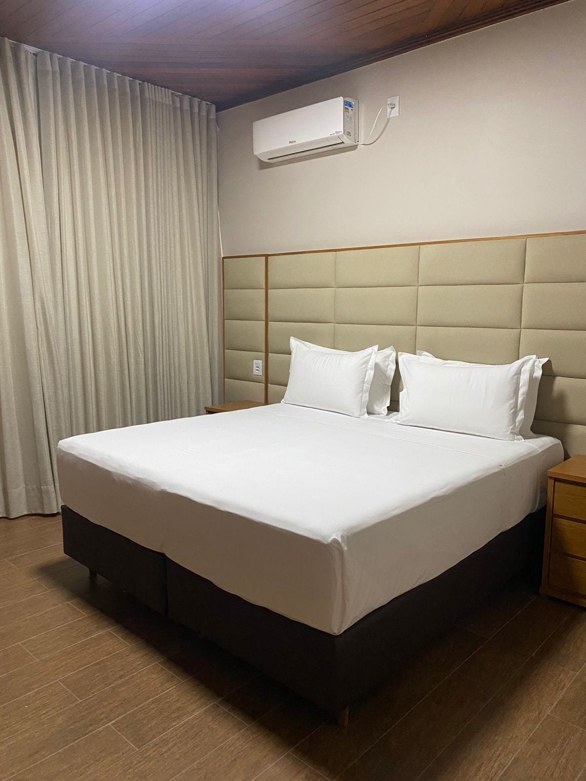 Une chambre d’hôtel avec un grand lit, une table de chevet en bois, un climatiseur fixé au mur et une fenêtre avec des rideaux.