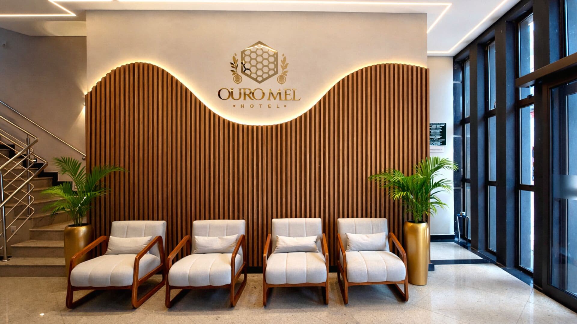 Uma área de recepção de hotel com design moderno, apresentando um logotipo acima de uma parede revestida de madeira e um espaço de estar com cadeiras e plantas em vasos.