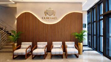 Uma área de recepção de hotel com design moderno, apresentando um logotipo acima de uma parede revestida de madeira e um espaço de estar com cadeiras e plantas em vasos.