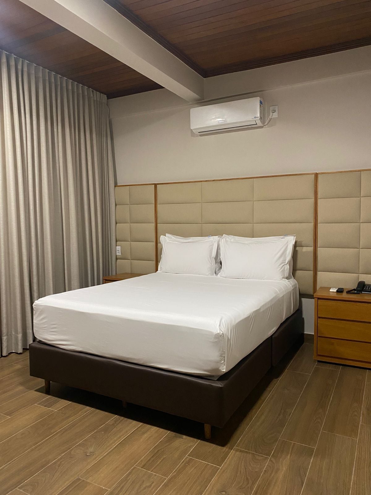 A hotel room with a bed, a nightstand, an air conditioning unit, and wooden flooring.
