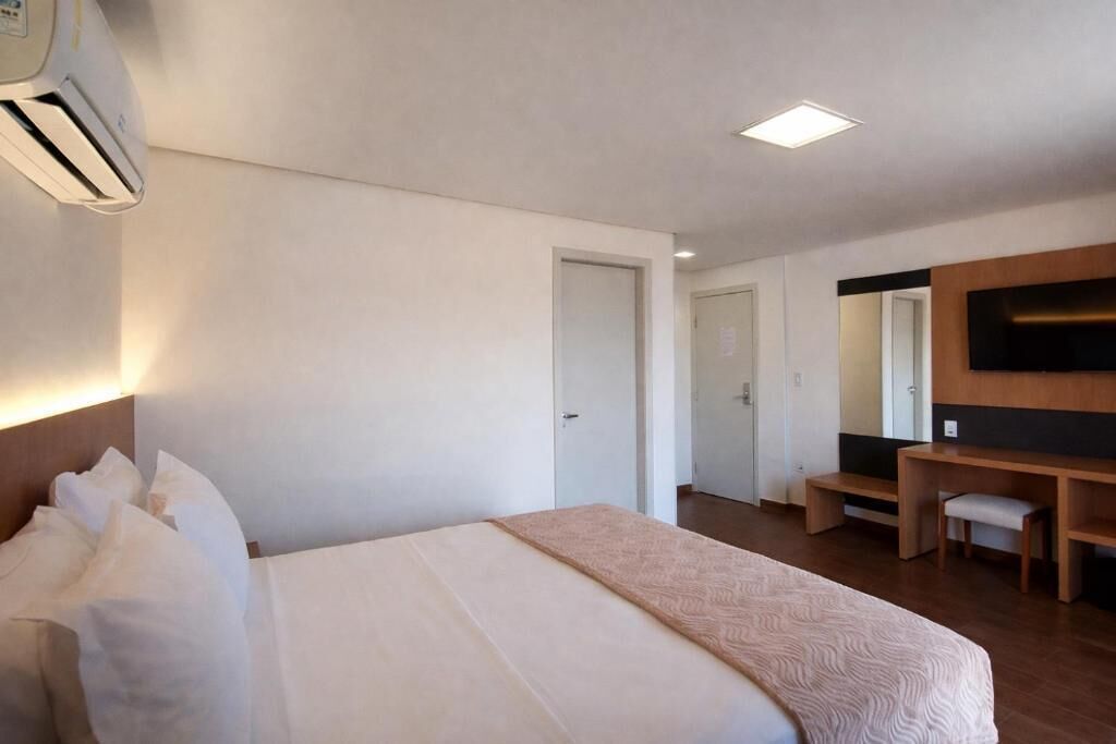 A hotel room with a bed, a desk, a chair, a TV, and an air conditioning unit.