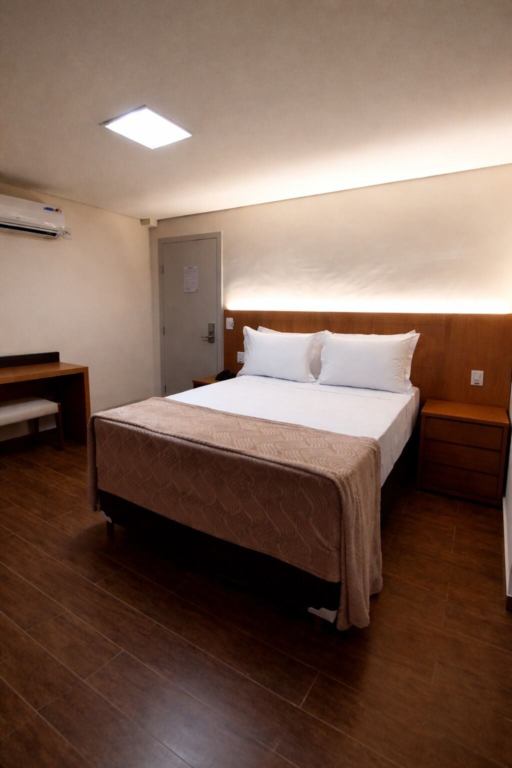 A hotel room with a bed, a bench, a bedside table, a nightstand, and a wall-mounted air conditioner.