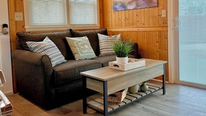 A cozy room with a wooden ceiling, a sofa with patterned cushions, a coffee table with a plant, and a painting on the wall.